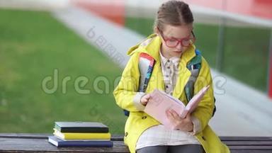 一个穿着黄色夹克和眼镜的优秀学生坐在学校附近的长凳上看书。 回到学校